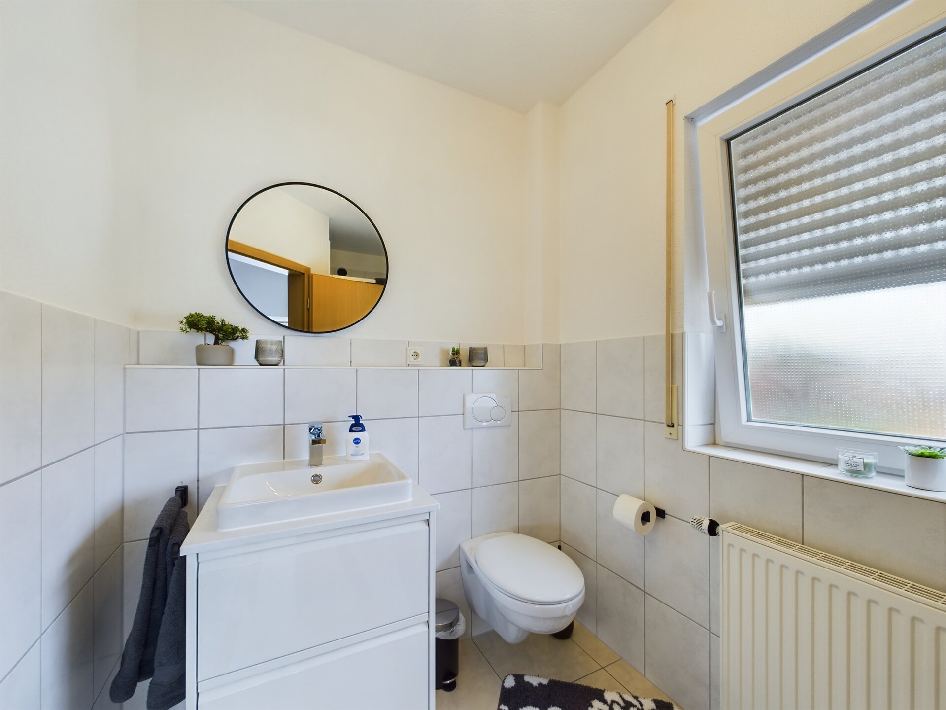 Modern bathroom with white tiles, a small sink, and a window with blinds near Ramstein Air Base.